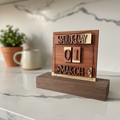 A wooden calendar block displaying Saturday, 01 March, styled on a dark wood base with a small potted succulent and a white vase in the background on a faux fur surface.
