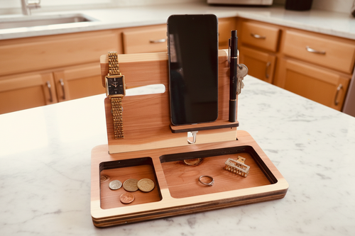 A wooden multi-functional desk organizer tray holding a smartphone, pen, watch, coins, ring, and small accessory, styled on a white faux fur surface.