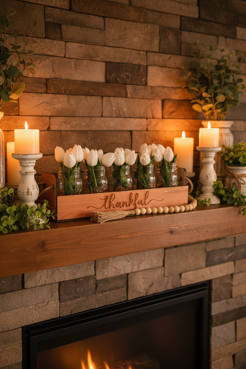 A wooden decorative tray engraved with the word 'thankful' holds several small clear glass jars filled with white tulips. The tray sits on a white faux-fur surface with a natural wood bead garland draped across the front.