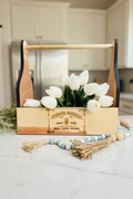 A wooden flower market caddy basket with a natural wood handle and sides, black inner trim, and a front label engraved with 'Flower Market Seeds - Stems - Blooms'. White tulips are arranged inside the basket, which sits on a white faux fur surface with a blue and beige beaded tassel garland in the foreground.