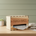 A wooden box labeled 'TRASH BAGS' with a white plastic trash bag being dispensed from the front opening, set on a white faux-fur surface against a light gray background.
