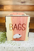 A wooden tissue bag dispenser box with a light natural wood top and sides, a cedar wood grain center panel, and the word 'BAGS' cut out on the front. A plastic grocery bag is visible in the opening, and the box is styled on a white textured surface with green leaves at the side.