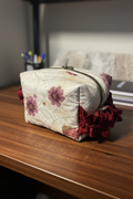 A small zippered cosmetic pouch with a cream-colored fabric body, embroidered floral and scroll patterns, purple flowers, and a dark red ruffled trim at the bottom, resting on a wooden surface.