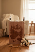 A rectangular wooden wall art piece with a laser-engraved mountain lake cabin landscape, displayed upright against a dark gray wall with decorative tulips and beads at the base.